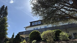 Une maison à deux étages, dotée d’un toit en pente, d’un balcon et d’un jardin agrémenté de diverses plantes et arbustes.