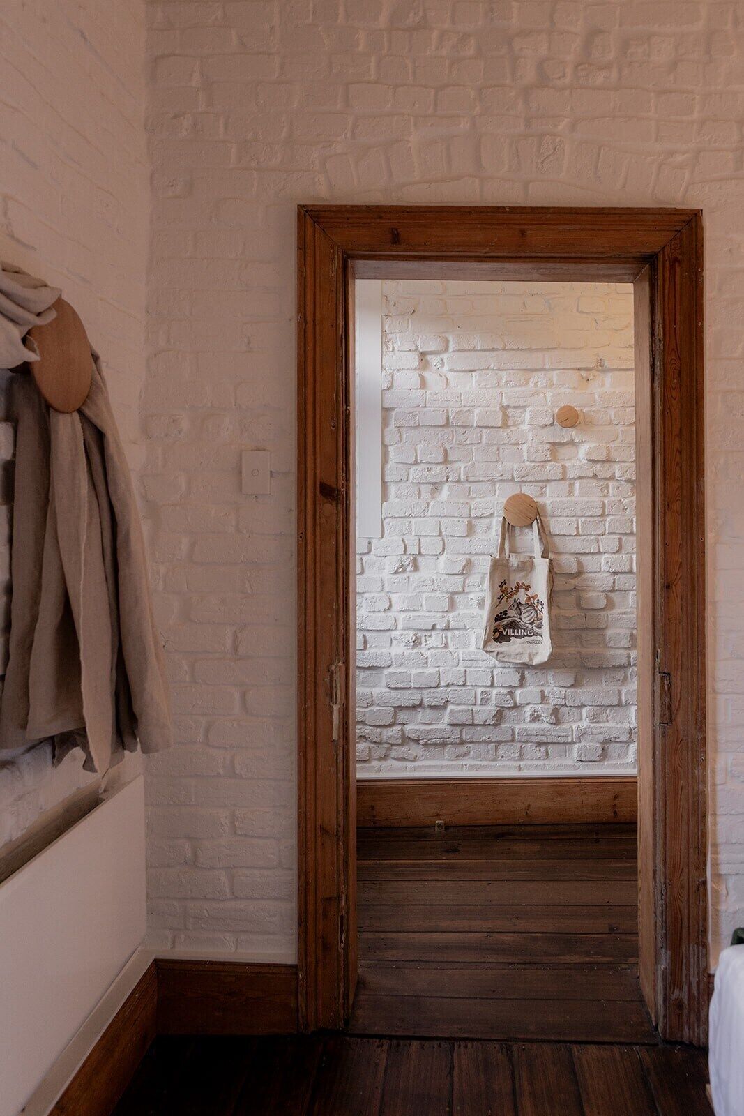 A narrow hallway with a white brick wall and a wooden door. A towel hangs on the wall, and a wooden bench is visible at the end of the hallway.