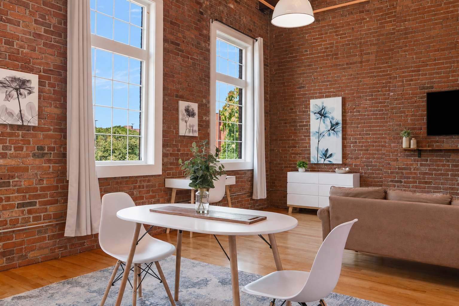 A modern living room with a brick wall, a round dining table, white chairs, a sofa, and artwork.