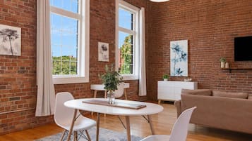 A modern living room with a brick wall, a round dining table, white chairs, a sofa, and artwork.