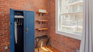 A room with a brick wall, a blue wardrobe, a rack with hangers, a bench, and a window with a view of buildings. - Jennifer’s Penthouse Loft | State House City Views (Providence)