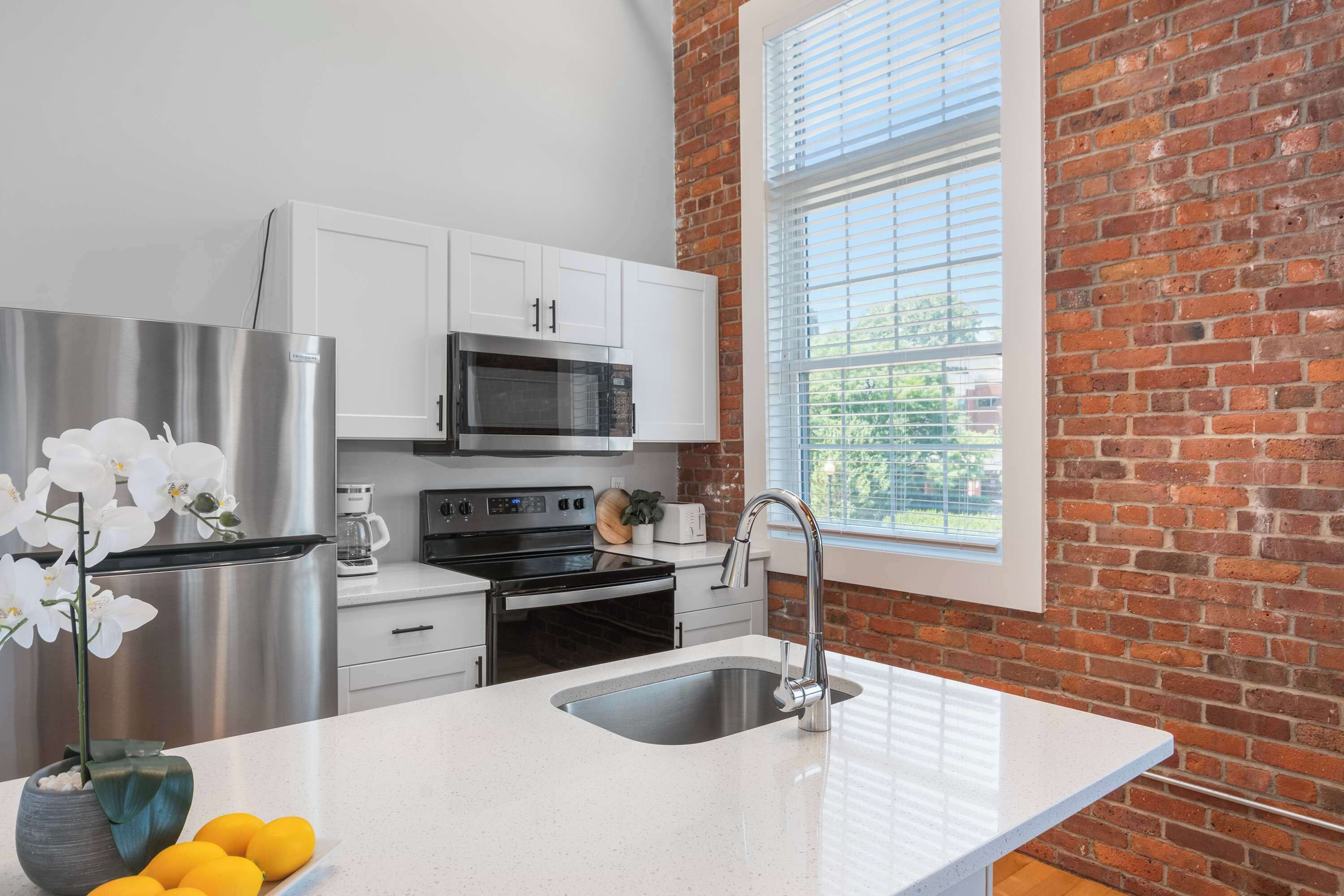 A modern kitchen with white cabinetry, stainless steel appliances, a large window with blinds, and a countertop with a sink and a vase of flowers.