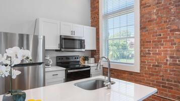 A modern kitchen with white cabinetry, stainless steel appliances, a large window with blinds, and a countertop with a sink and a vase of flowers.