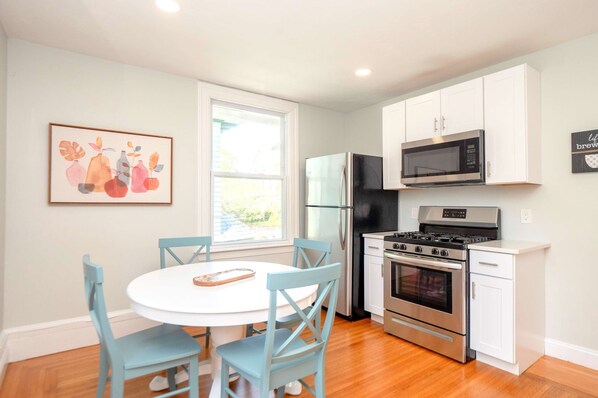 A modern kitchen with white cabinets, a stainless steel stove, a microwave, and a refrigerator. There is a dining table with blue chairs and a window providing natural light.