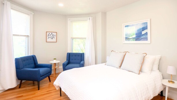 Une chambre à coucher avec un grand lit, deux fauteuils bleus, une petite table ronde et un tableau accroché au mur.