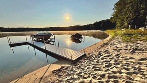 Hiekkainen ranta, josta ulottuu tyynen veden ylle puinen laituri, laiturissa kiinni oleva vene ja kirkas taivas, jossa aurinko on matalalla horisontissa.
