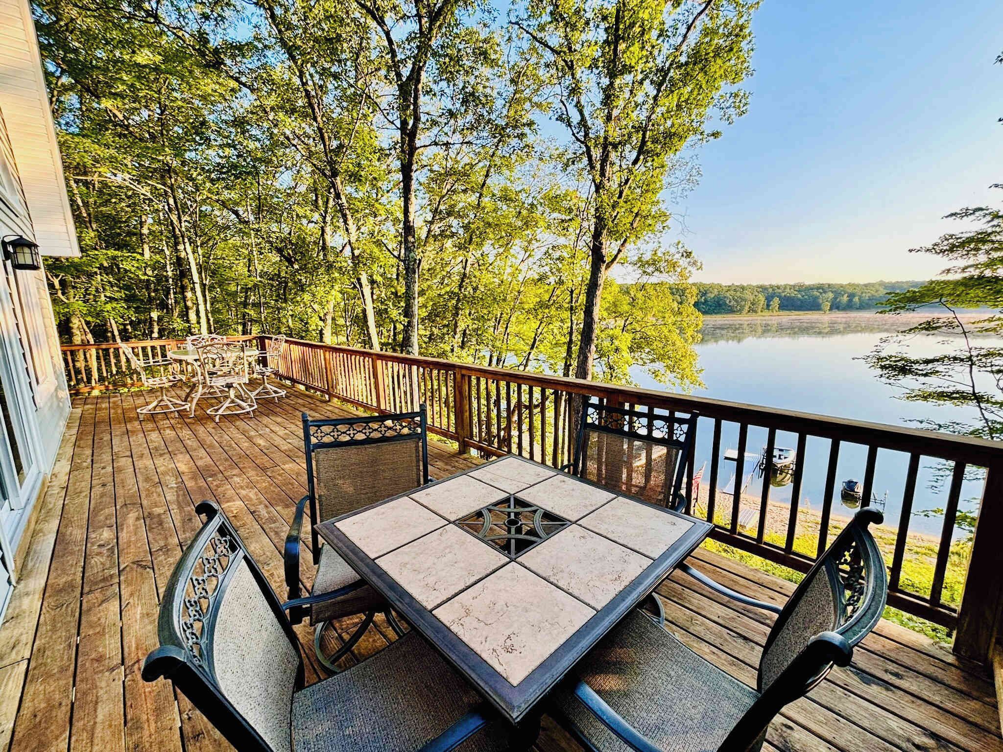 Une terrasse en bois avec une table et des chaises, donnant sur un lac et entourée d’arbres.