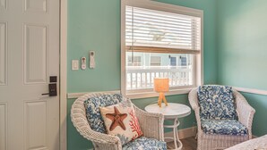 Ein Zimmer mit türkisfarbenen Wänden, einer weißen Tür, zwei Korbstühlen mit Blumenkissen, einem kleinen runden Tisch mit einer Lampe und einem Fenster mit Jalousien.