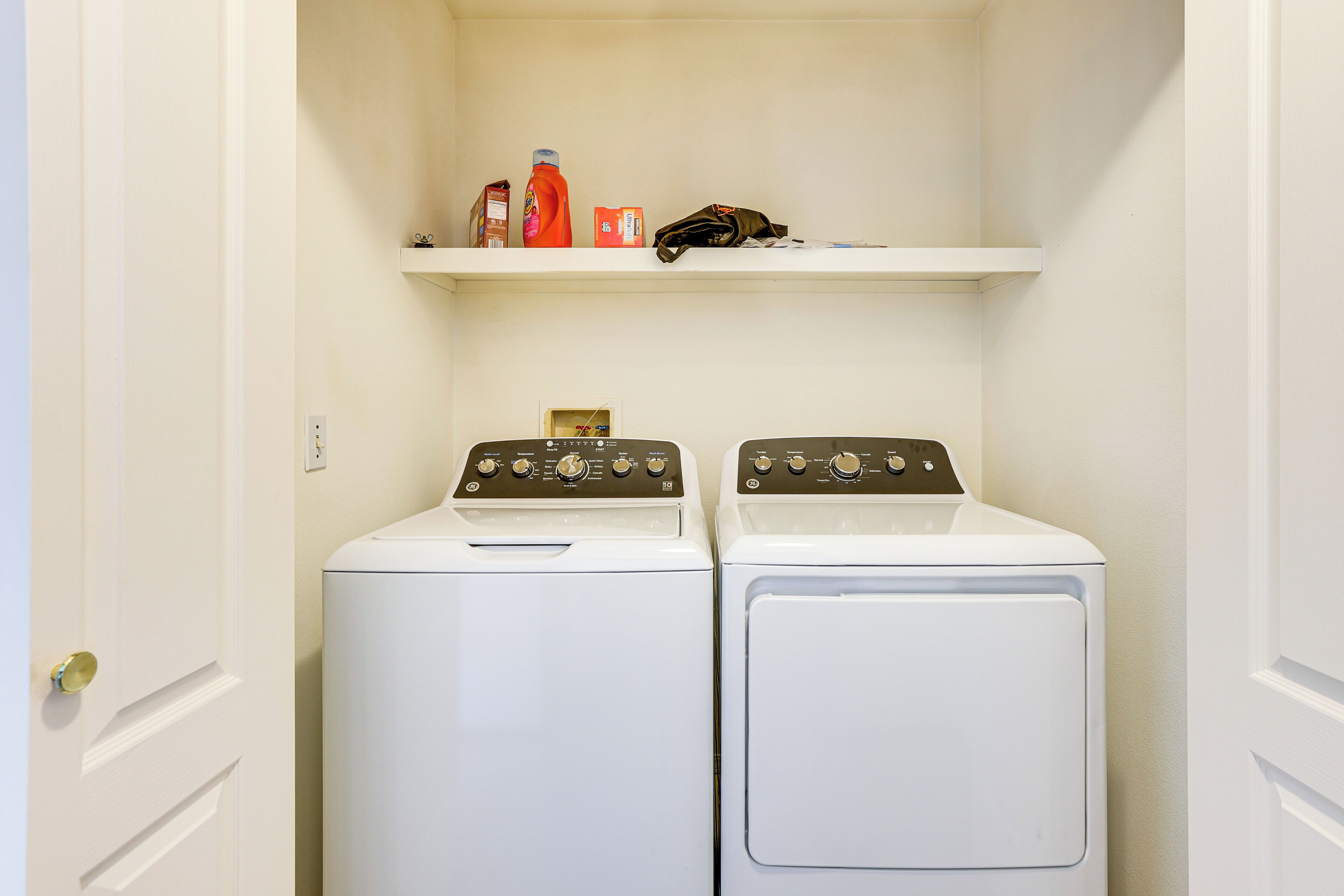 A laundry room with two white front-loading washers and a shelf above containing cleaning supplies.