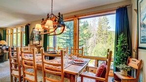A dining area with a wooden table and chairs, a chandelier, and a view of trees through large windows. - Gem in Old Town Winter Park: Walk to Ski Lifts! (Winter Park)