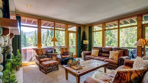 A spacious living room with a large wooden coffee table, leather sofas, and a view of a forest through large windows. - Gem in Old Town Winter Park: Walk to Ski Lifts! (Winter Park)
