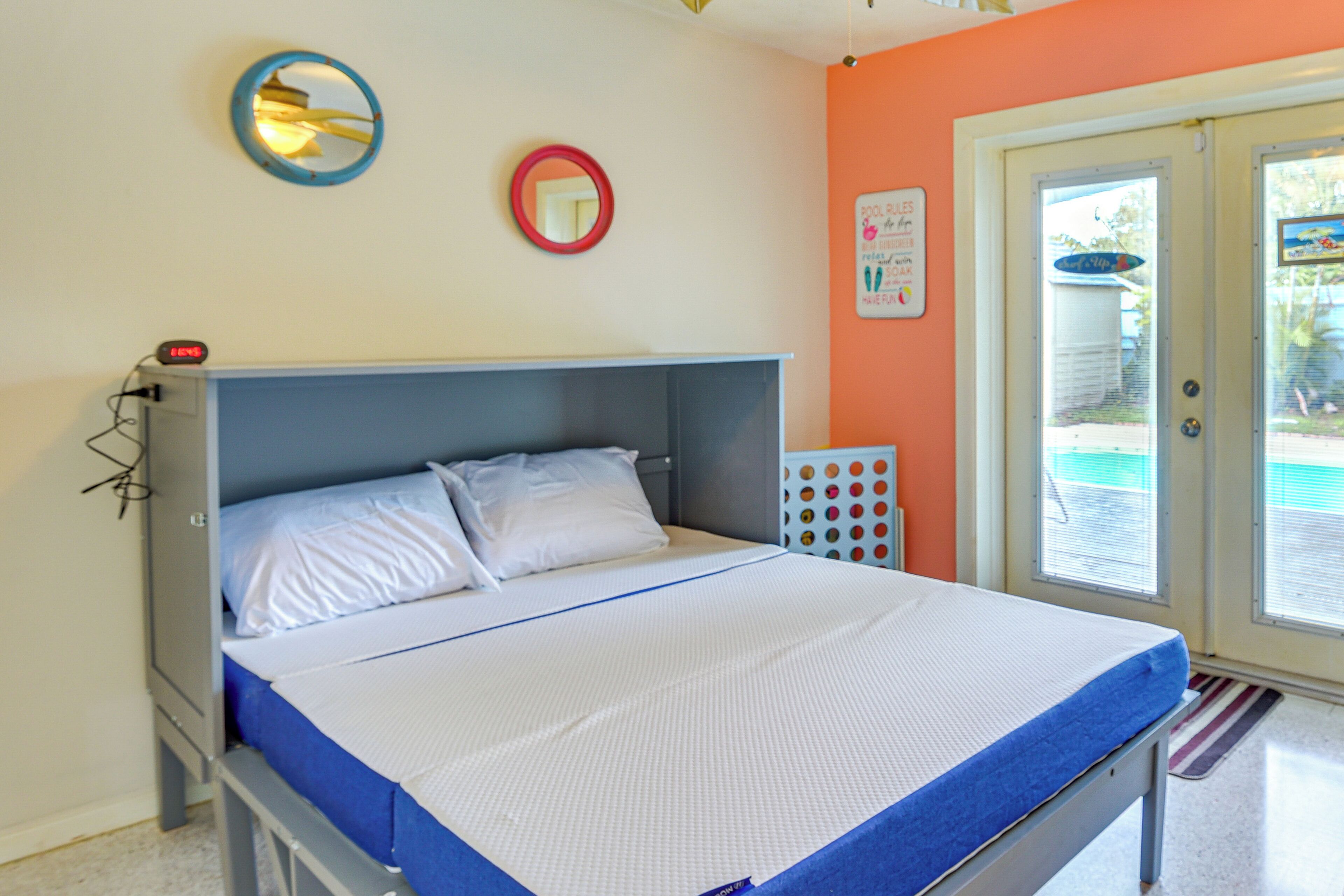 A single bed with a grey headboard, white bedding, and a blue accent at the foot of the bed. There is a small round mirror on the wall above the bed and a sign on the door.