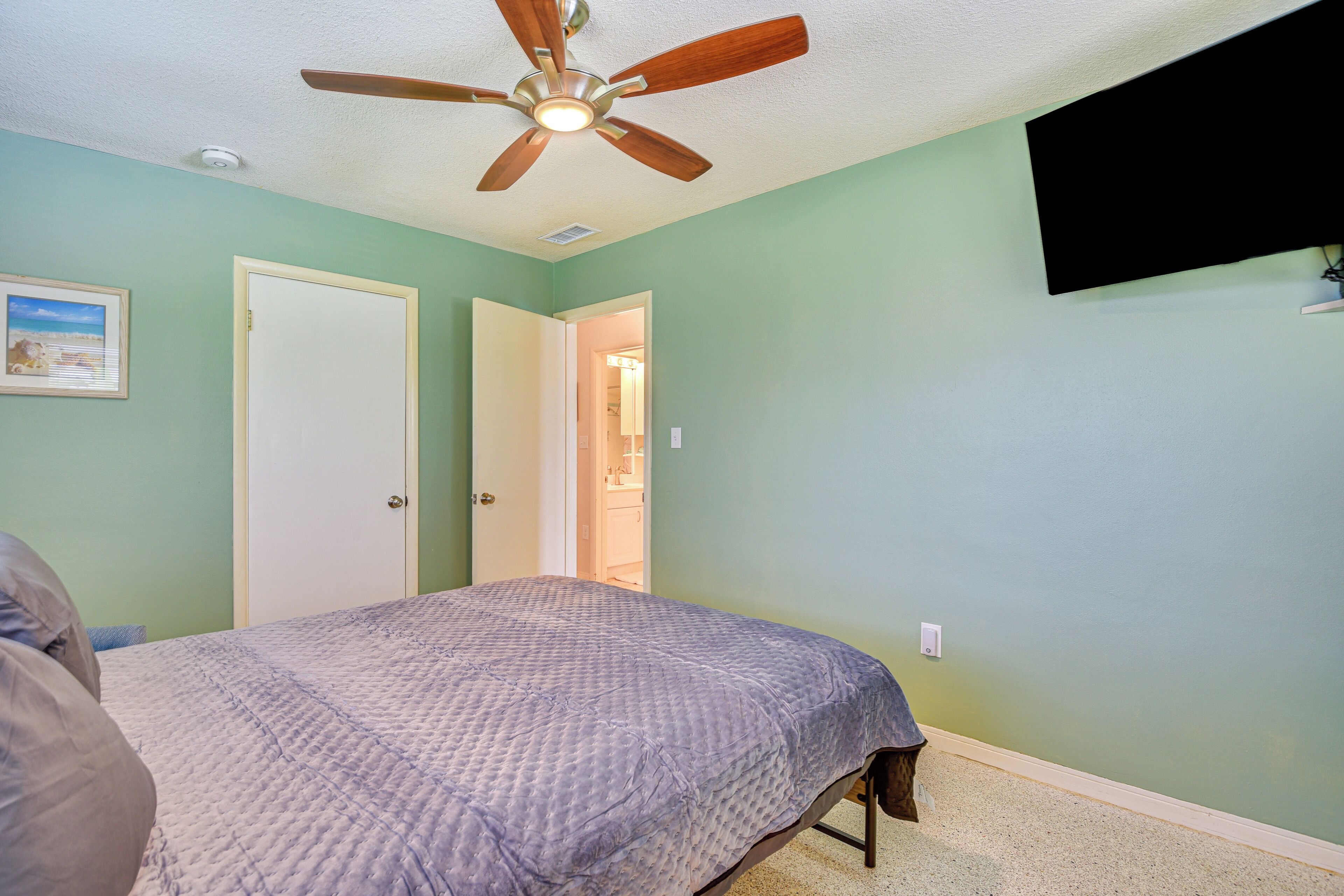 A bedroom with a bed, a ceiling fan, a door, and a wall-mounted TV.