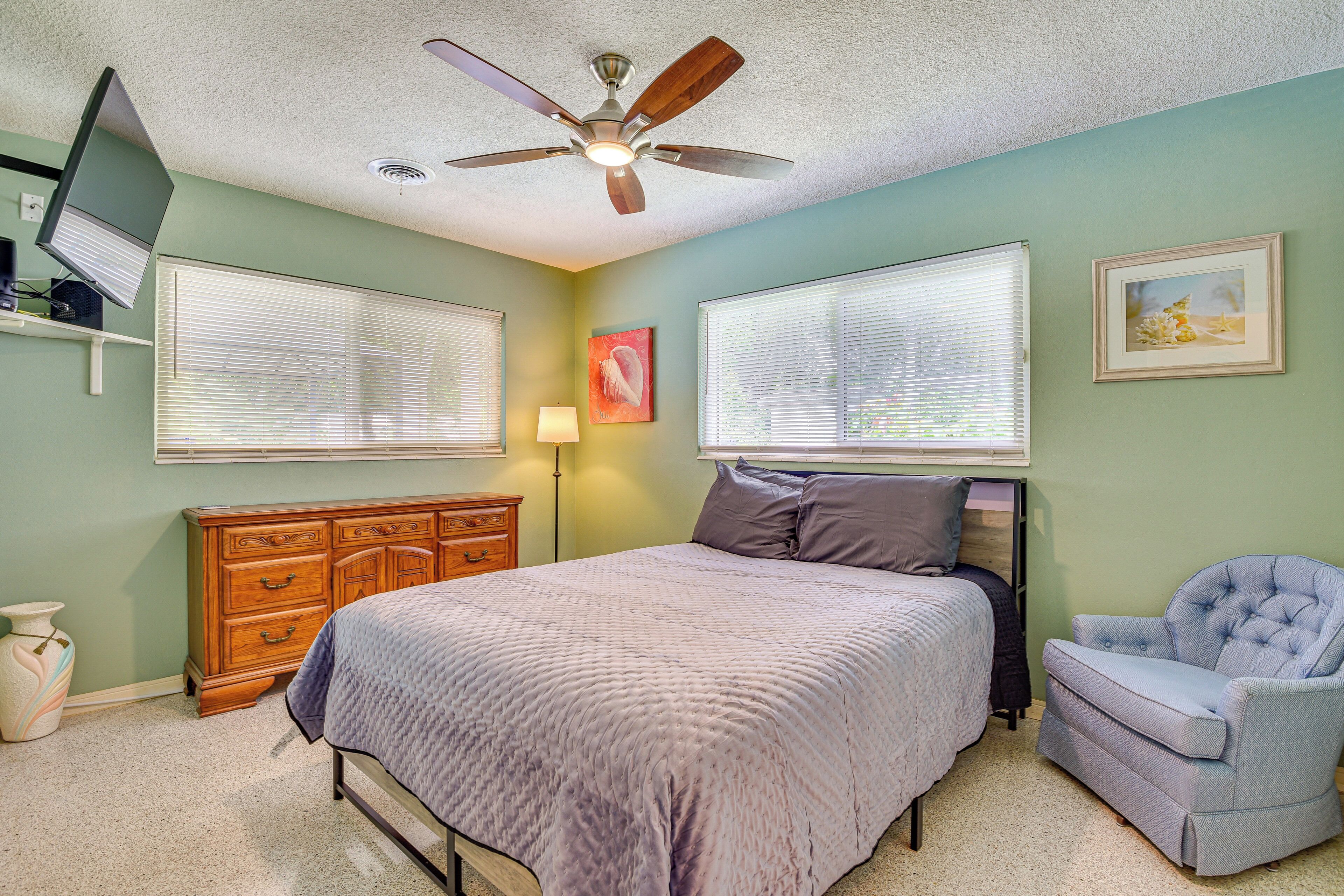A bedroom with a bed, dresser, chair, ceiling fan, and two windows with blinds.