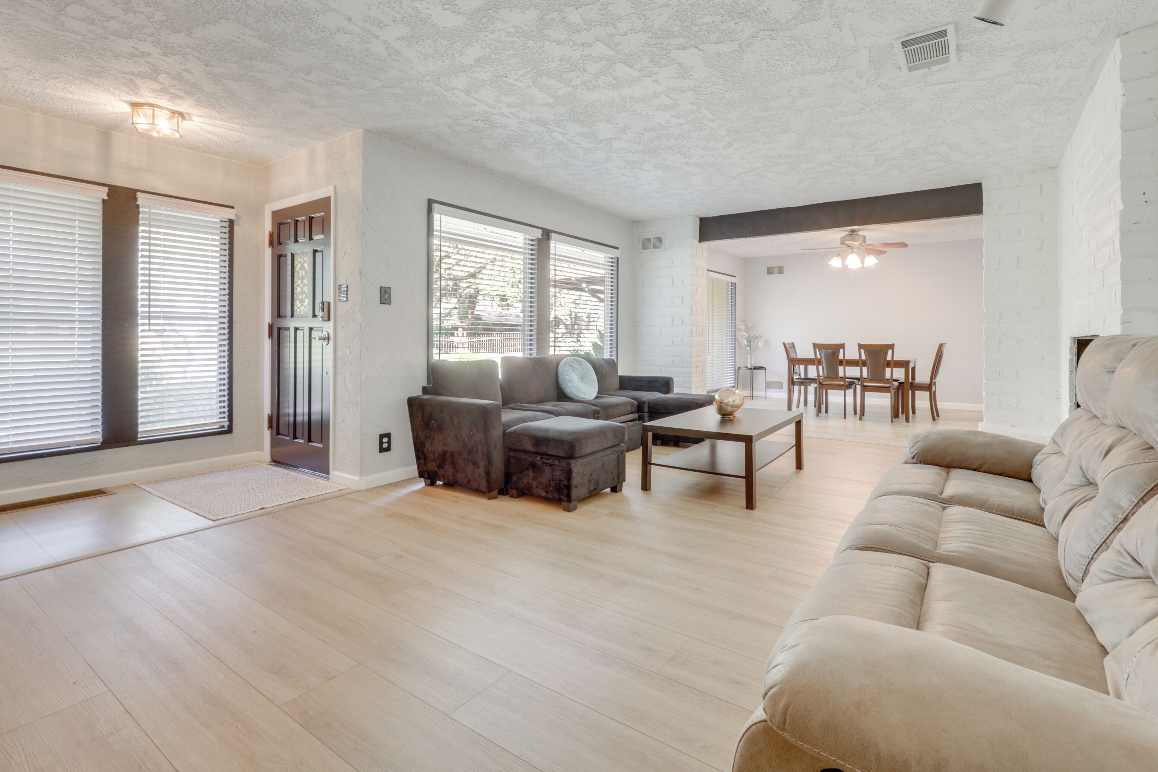 A spacious living room with a beige sofa, a dark-colored armchair, and a small table. There is a dining area with a wooden table and chairs. The room has large windows with blinds, a white brick wall, and a ceiling fan.