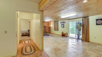 A spacious living area with a wooden ceiling, a large window with curtains, a dartboard, and a bookshelf.