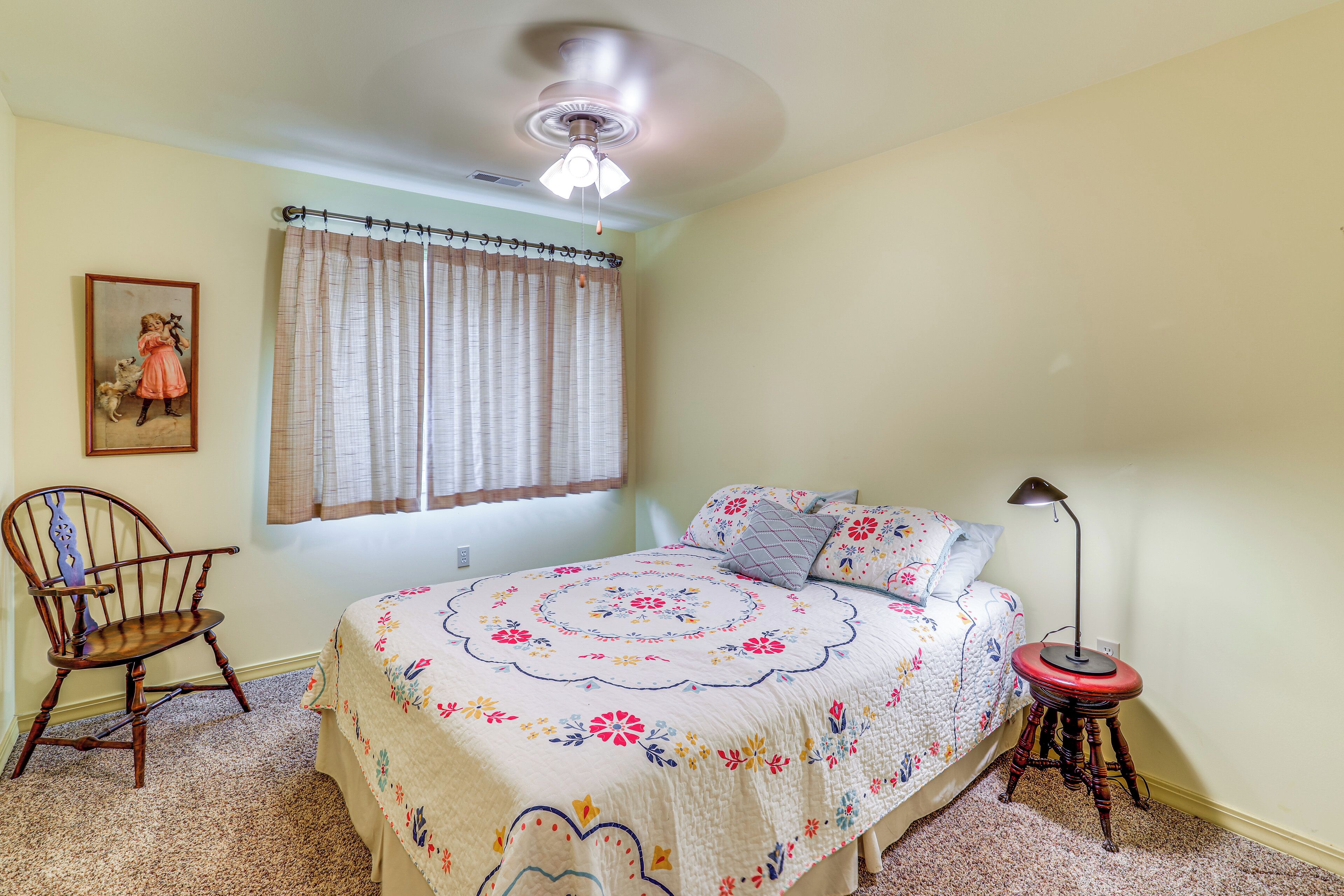 A bedroom with a bed, a chair, a lamp, a window with curtains, and a ceiling fan.