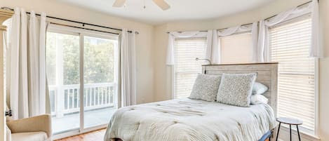 A bedroom with a bed, a ceiling fan, a chair, and a balcony with a view of trees.