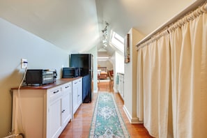 Um corredor estreito com uma área de cozinha à esquerda, com armários brancos, bancada com torradeira e micro-ondas, e uma geladeira preta. Um tapete azul estampado percorre o corredor, levando a uma sala de jantar com mesa e cadeiras de madeira.