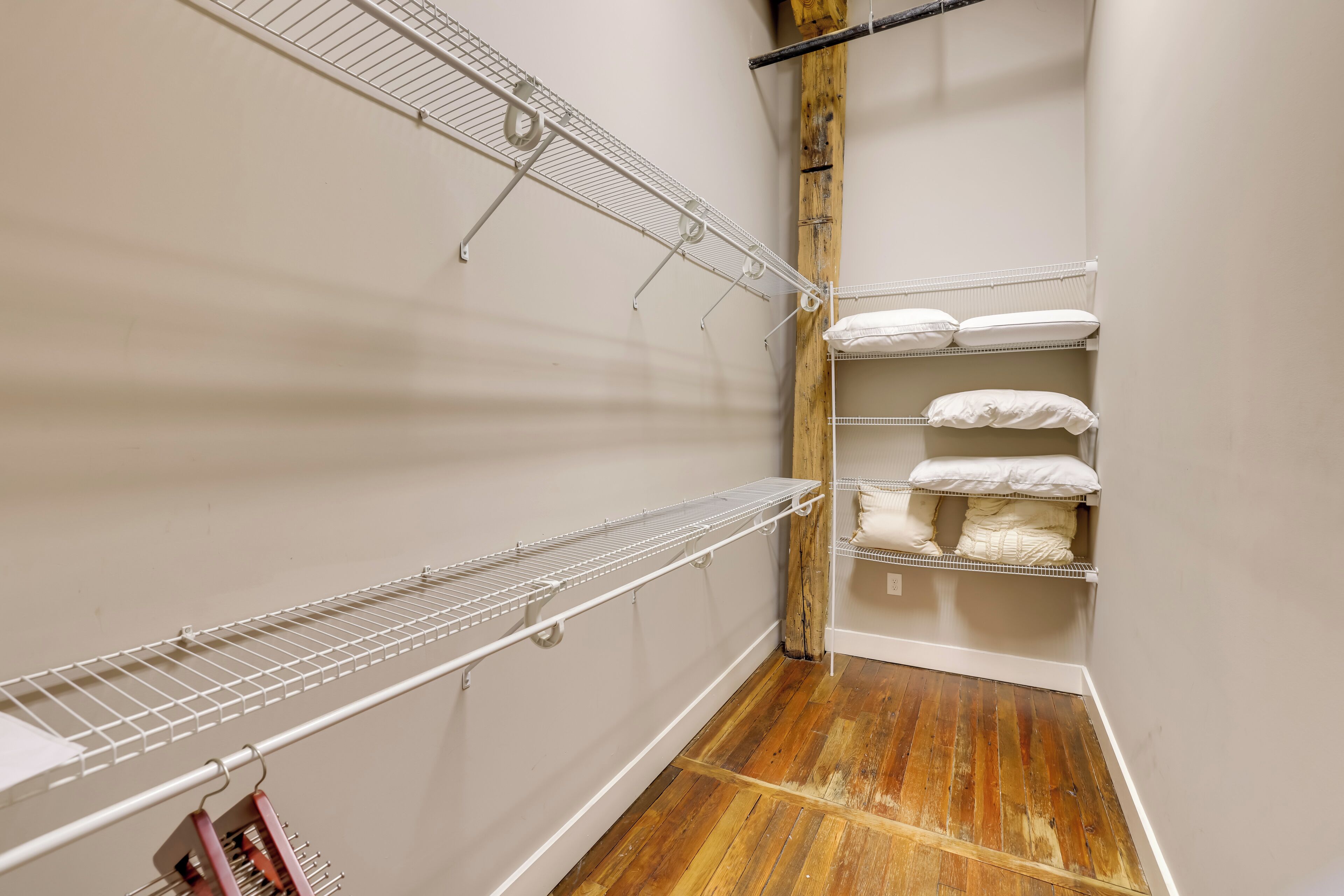 A narrow hallway with wooden flooring and white walls. There are two white wire shelves on the left and a wooden beam on the right.