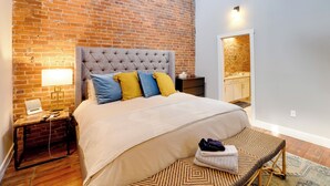 Une chambre avec un mur en briques, une tête de lit capitonnée, un lit recouvert d’une couette beige, une table de chevet avec une lampe et un petit banc avec des serviettes.