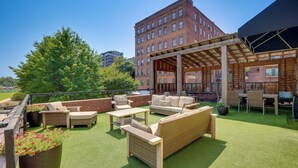 Un espace extérieur aménagé en patio, avec des sièges et une pergola, entouré d’un bâtiment en briques et de verdure.