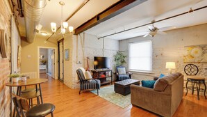 A spacious living room with a wooden floor, a sofa, a coffee table, and a TV. There is a brick wall, a ceiling fan, and a window with blinds. - Dtwn Lynchburg Loft w/ Shared Deck: Walk to River (Lynchburg)