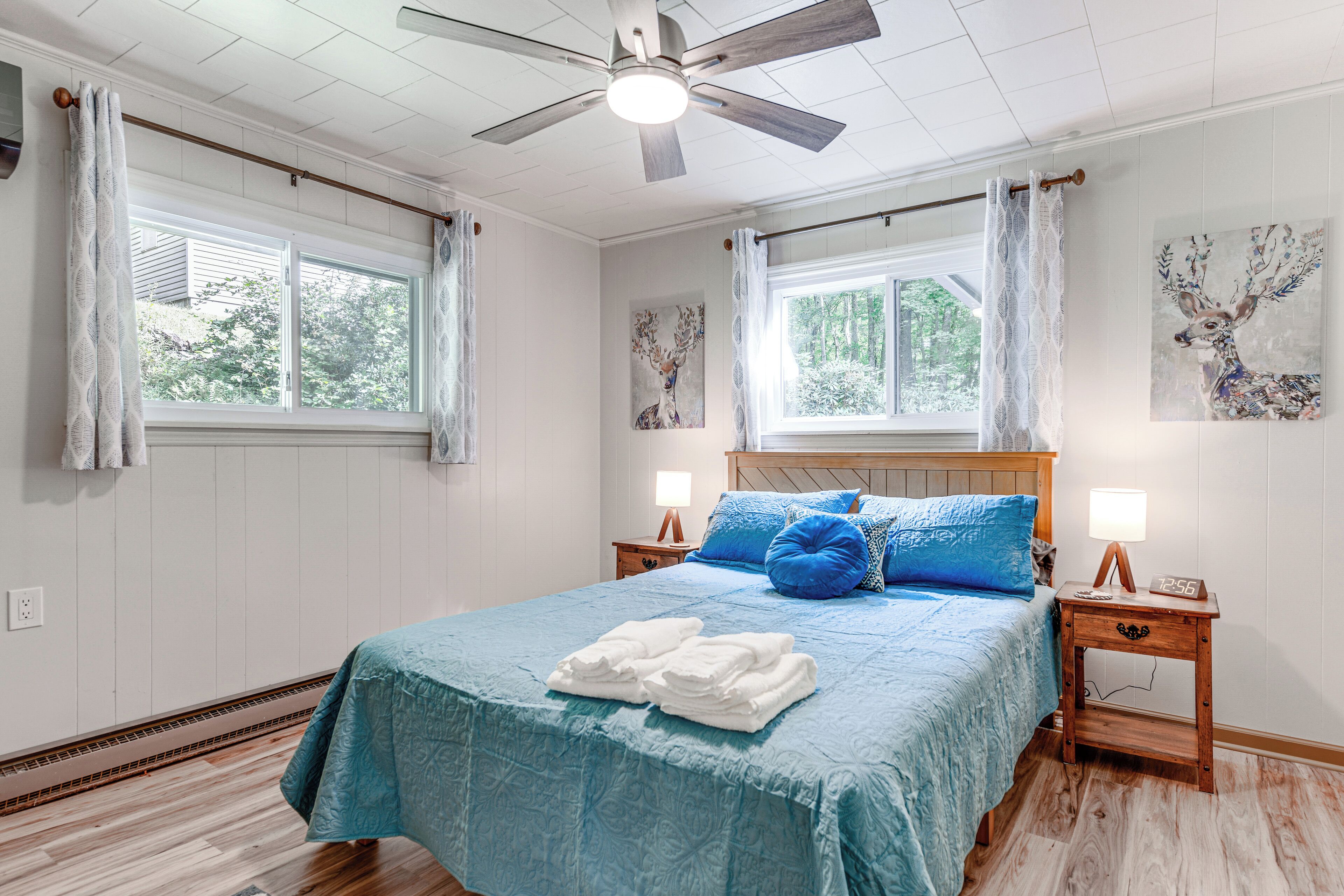 A bedroom with a large bed, two bedside tables with lamps, a ceiling fan, and two windows with curtains.