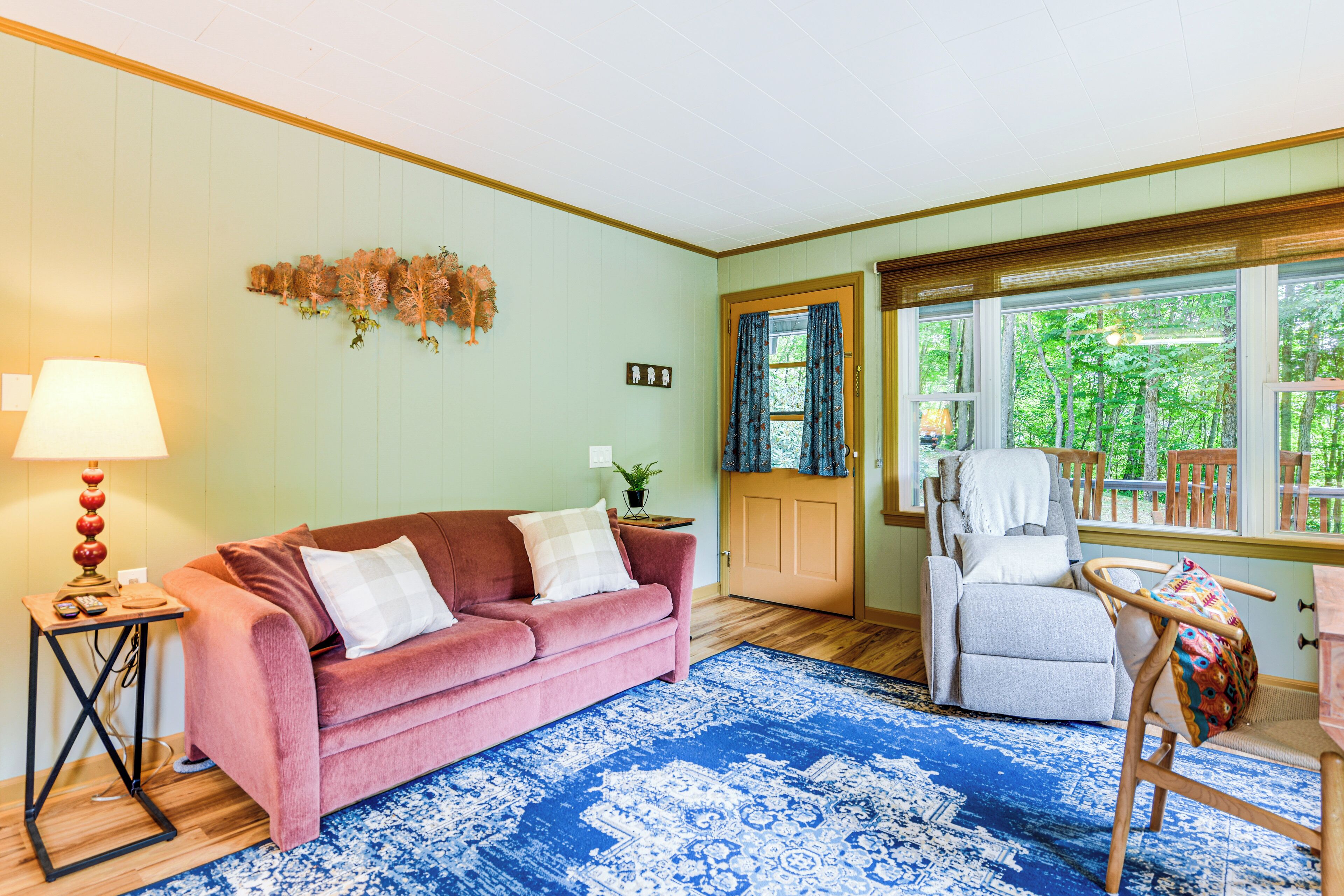 A living room with a pink sofa, a blue patterned rug, and a wooden floor. There is a lamp on a side table, a chair, and a window with curtains.