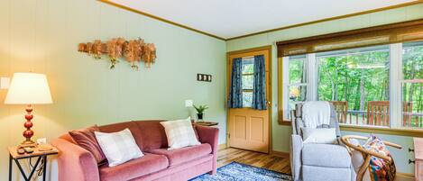 A living room with a pink sofa, a blue patterned rug, and a wooden floor. There is a lamp on a side table, a chair, and a window with curtains.