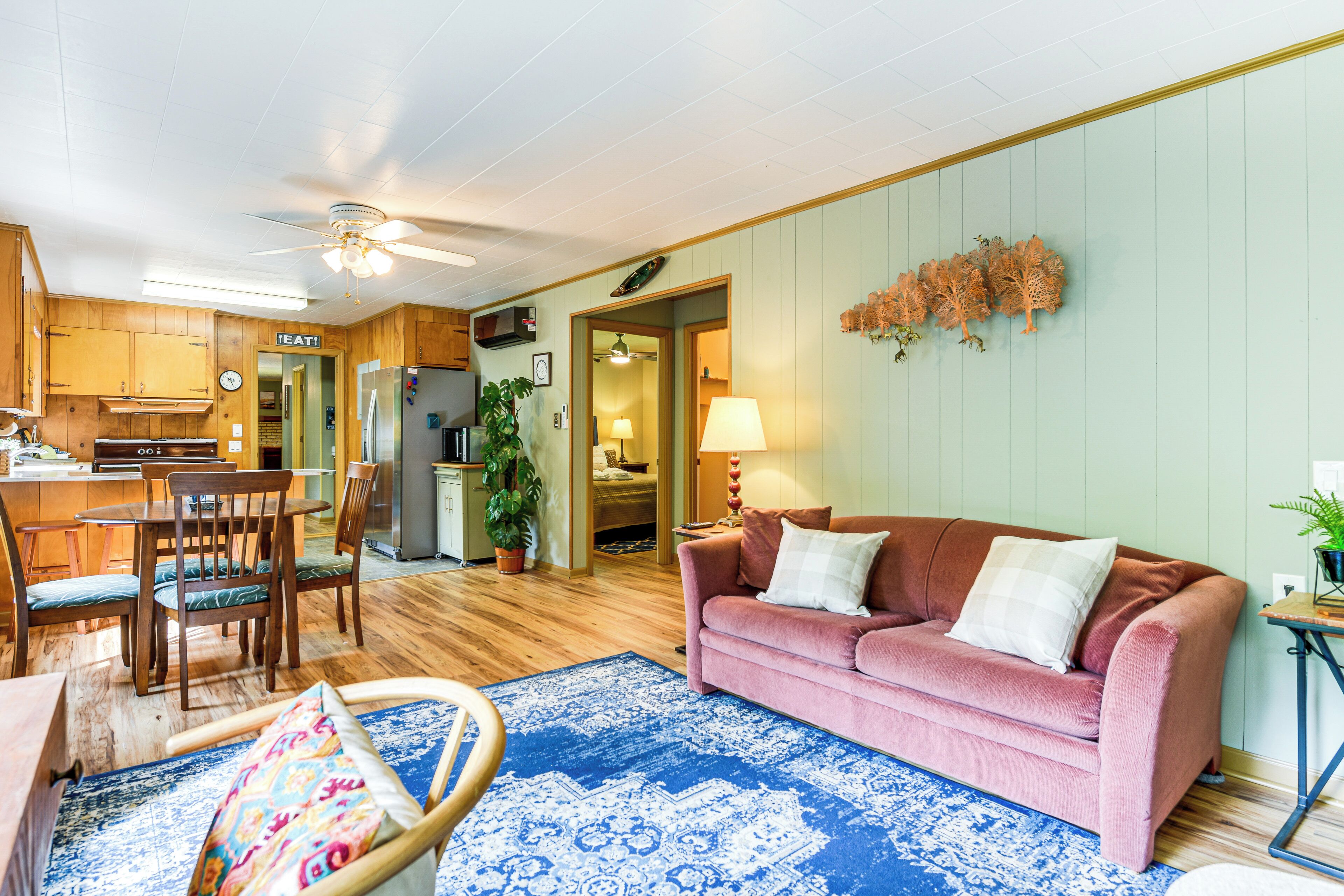 A cozy living room with a pink sofa, wooden dining table, and chairs. A blue patterned rug is on the floor.