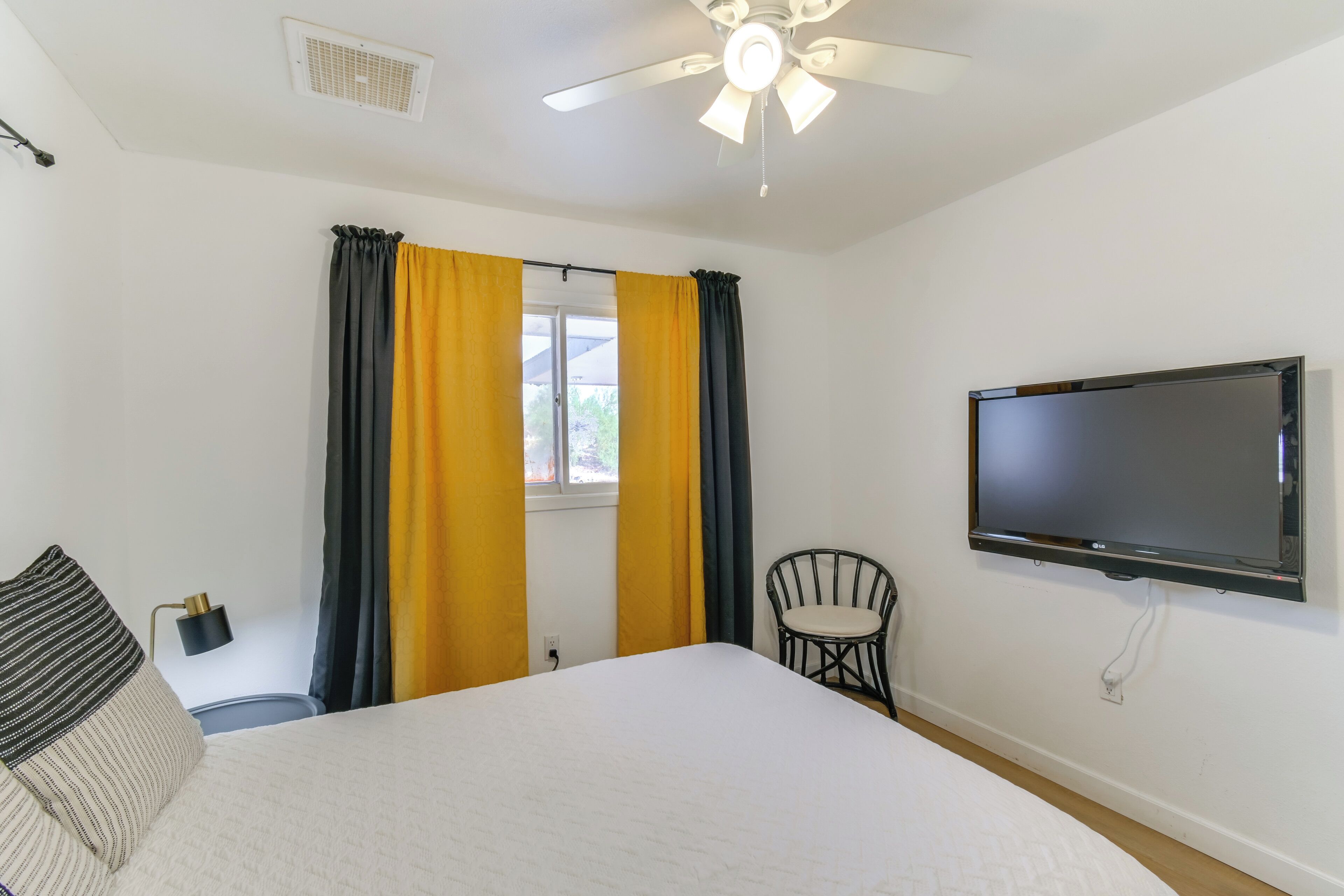 Une chambre d’hôtel avec un lit, un téléviseur fixé au mur, un ventilateur de plafond et une fenêtre avec des rideaux.