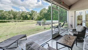 Eine Terrasse mit Gartenmöbeln, eine Glastür zu einer Rasenfläche und Blick auf Bäume und einen See.