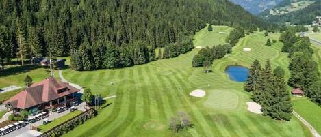 Vista aérea de um campo de golfe com um clube, rodeado por floresta densa e colinas.