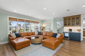 A living room with a sectional sofa, a coffee table, and a kitchen area with cabinets and a stove. - The Sea Suite: Lovely Sugar Sands Home- Priv. Pool, Sleeps 18, Game & Movie Room (Mexico Beach)