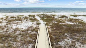 Ein hölzerner Steg führt zu einem Strand mit weißem Sand und spärlicher Vegetation.