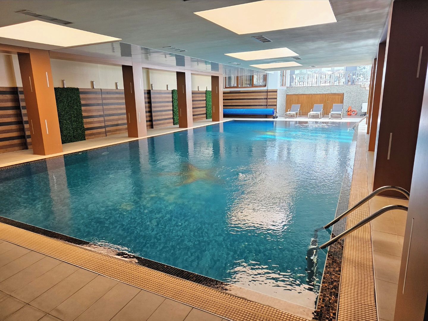 An indoor swimming pool with clear water, surrounded by walls with a striped pattern and green plants. There are lounge chairs and a lifebuoy visible.
