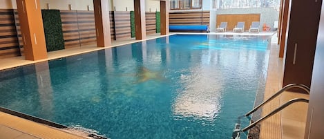 Una piscina interior con agua cristalina, rodeada de paredes con diseño a rayas y plantas verdes. Se observan sillas de descanso y un flotador.