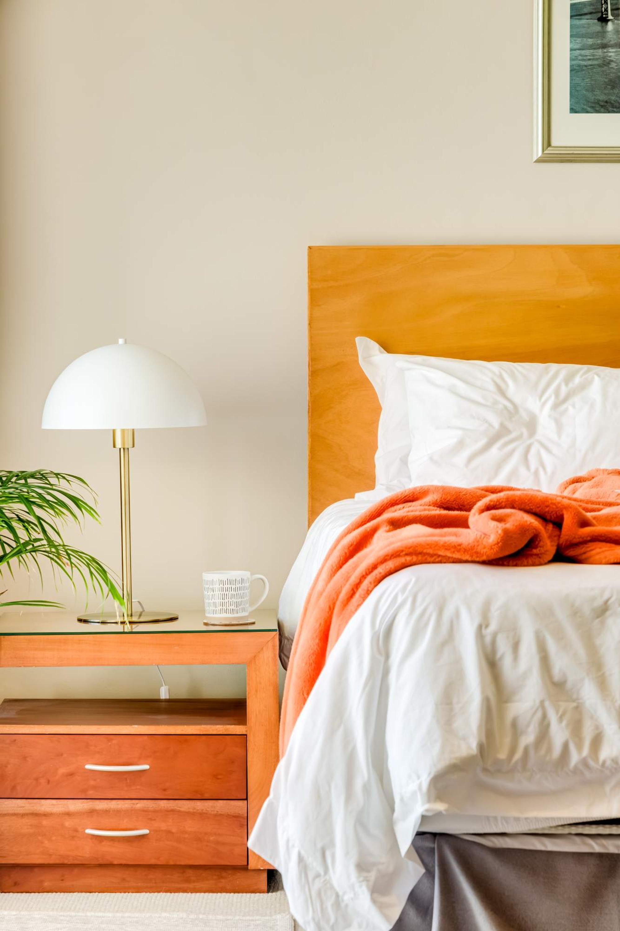 A neatly made bed with a white headboard and an orange blanket, a bedside table with a lamp, and a framed picture on the wall.