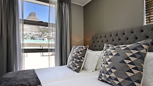 Hotelkamer met een groot bed, een grijs gestoffeerd hoofdeinde en uitzicht op een berg en gebouwen door het raam.