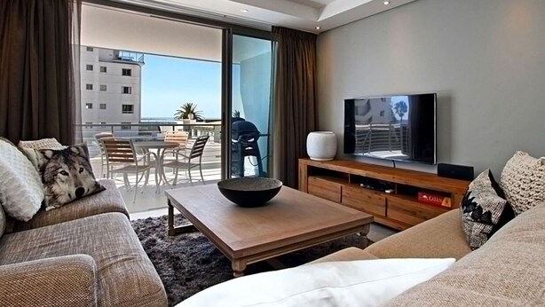 Een moderne woonkamer met een bank, een bijzettafeltje en een televisie. Er is een balkon met uitzicht op gebouwen en een tafel buiten.