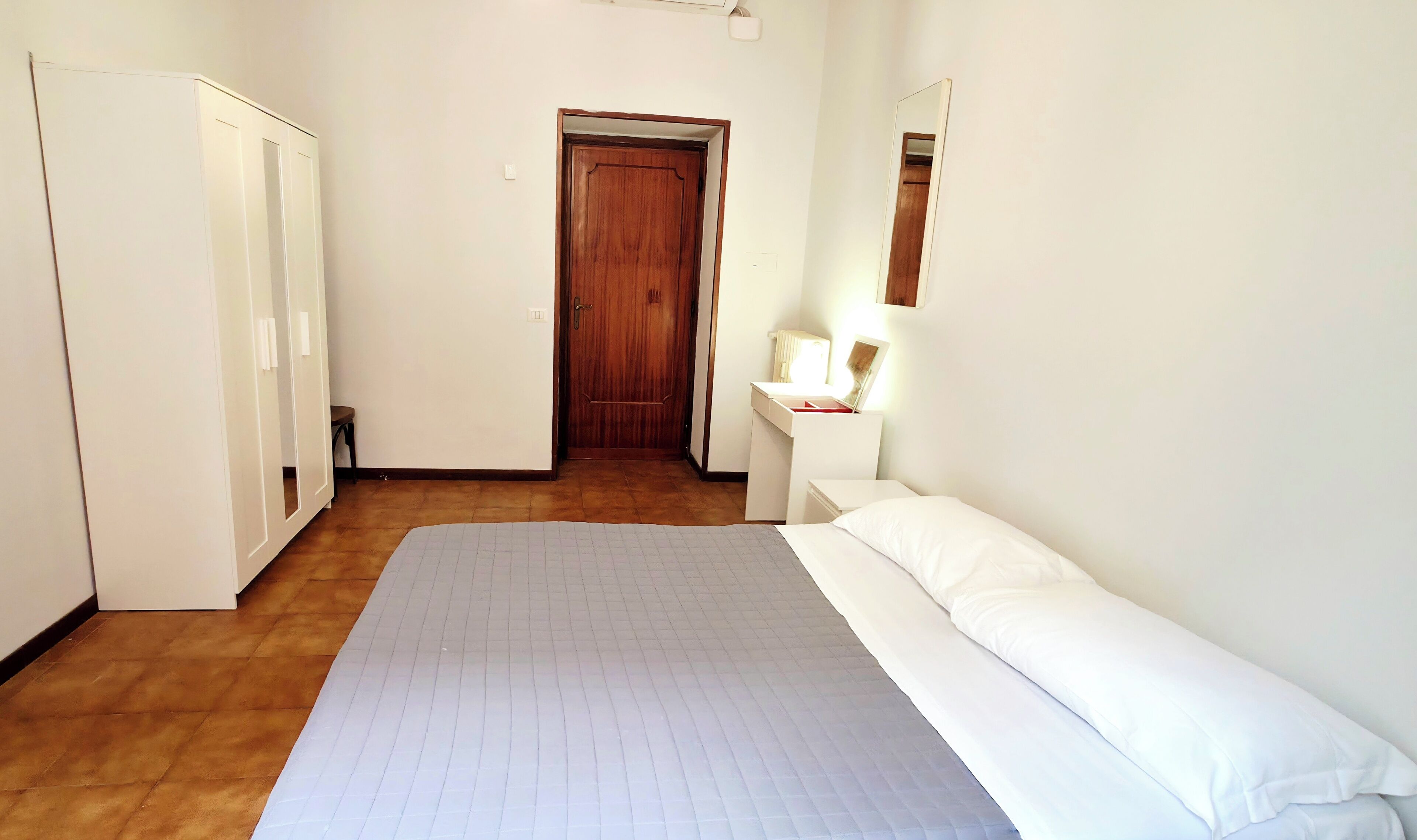 A bedroom with a bed, a nightstand, a mirror, a wardrobe, and a door leading to another room.