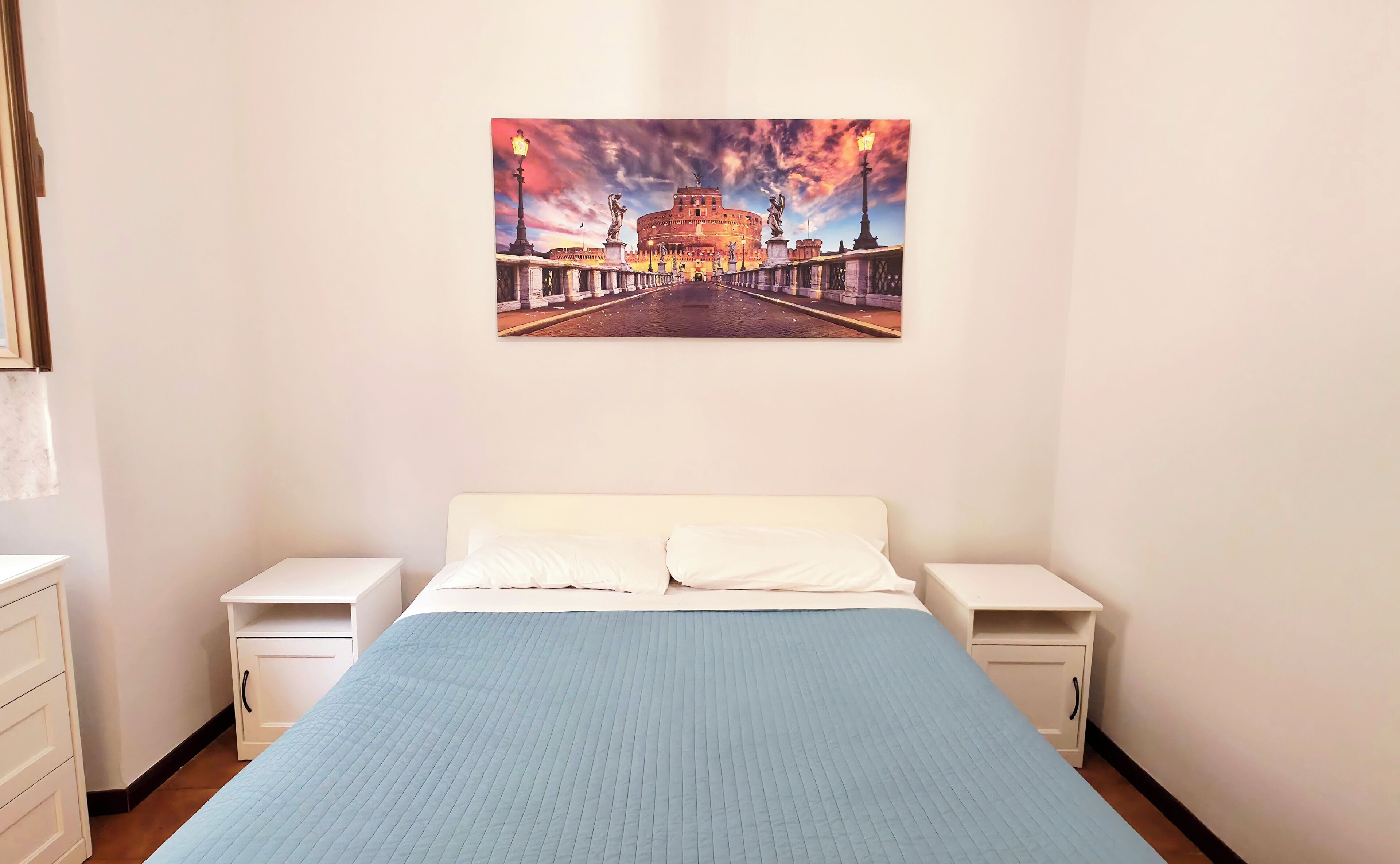 Un dormitorio con una cama, dos mesitas de noche, una lámpara de noche, un cuadro colgado en la pared de un edificio histórico y una ventana con persianas.