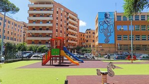 A playground with slides and a swing set, surrounded by multi-story buildings and parked cars. - Latini's Home (Rome)