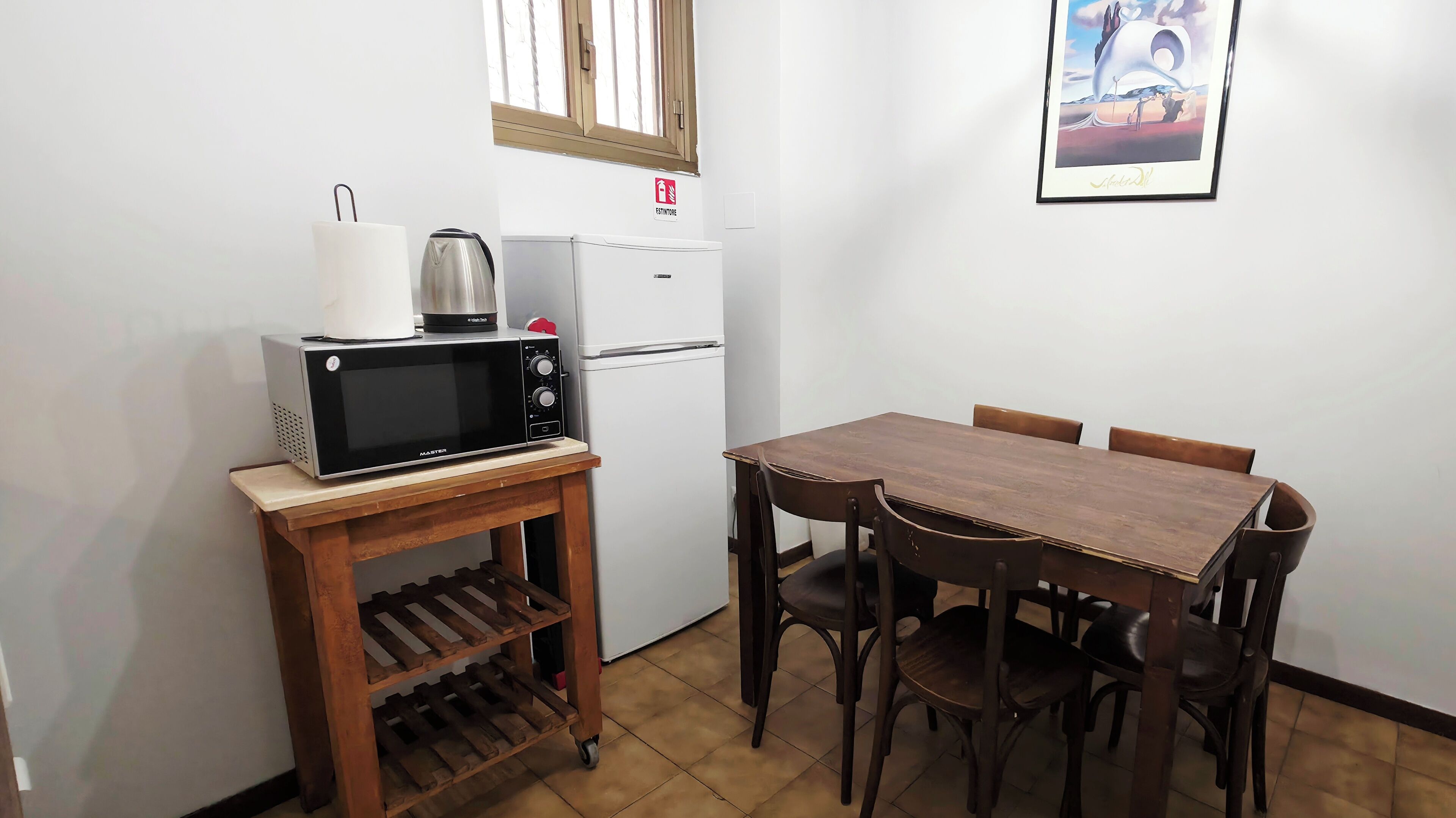 A compact kitchen with a microwave, kettle, and refrigerator, a wooden table with chairs, and a framed picture on the wall.