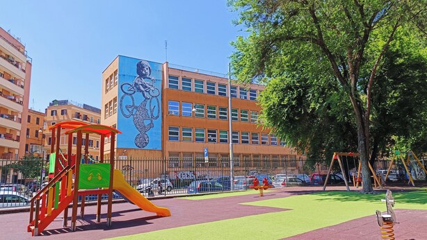 A playground with a slide and climbing structure, surrounded by a fenced area with a parking lot and buildings in the background. - Latini's Home (Rome)