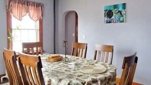 A dining area with a round table, four wooden chairs, a floral tablecloth, a window with curtains, and a painting on the wall.