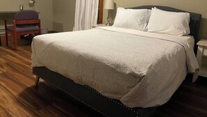 A neatly made bed with a dark headboard, a bedside lamp, and a chair in a room with wooden flooring and a window with white curtains.