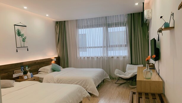 Et hotelværelse med to senge, en træsenggavl, et natbord med lampe, en stol, et skrivebord med en vase og et vindue med let gardin.