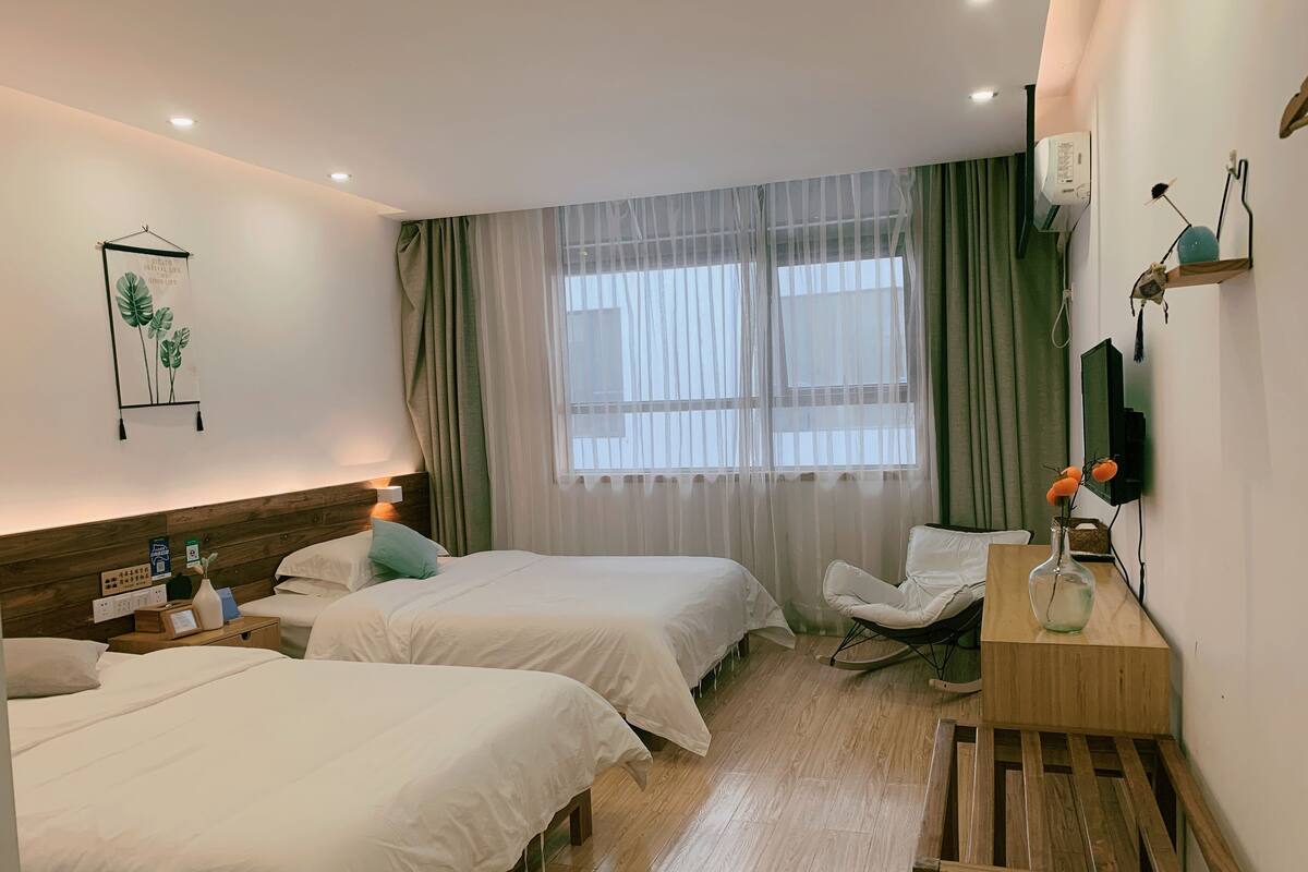 Habitación de hotel con dos camas, cabecera de madera, mesita de noche con lámpara, una silla, escritorio con un florero y ventana con cortinas traslúcidas.